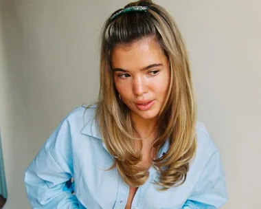 Woman with long, wavy blonde hair styled into a pouf, wearing a light blue shirt, looking to the side.