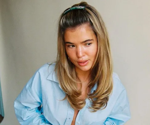 Woman with long, wavy blonde hair styled into a pouf, wearing a light blue shirt, looking to the side.