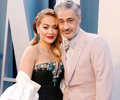 A couple poses at an event; she wears a floral black dress with diamond earrings, he dons a beige suit with a bow tie.