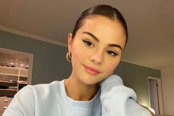 A person with sleek hairstyle, winged eyeliner, and hoop earrings, wearing a light blue sweatshirt, sitting indoors.