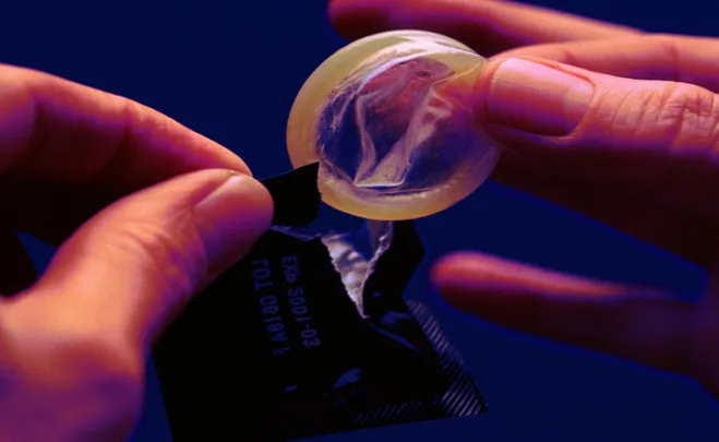 A person holding an opened condom wrapper and a condom against a blue background.