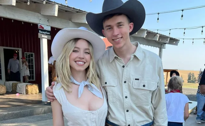 Two people dressed in cowboy hats and shirts smiling outdoors at a rustic venue.