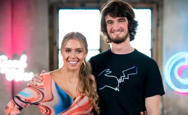 Woman in colorful dress and man in black shirt smiling indoors with neon signs in the background.