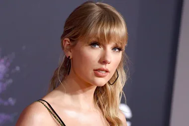 A woman with long blonde hair and bangs, wearing black earrings and a strappy top, posing in front of a blurred background.