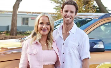 A smiling couple stands in front of a car, outdoors, with trees in the background.