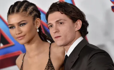 A man and a woman with braided hair pose at a formal event, standing side by side against a colorful backdrop.