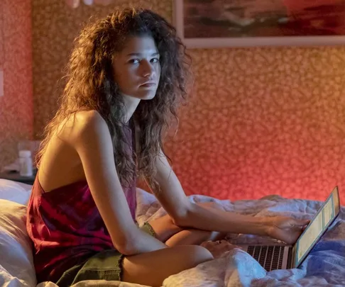 Girl with curly hair sitting on a bed, using a laptop in a warmly lit room.