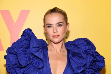 Zoey Deutch in a bold blue dress with dramatic ruffled shoulders at a "Not Okay" movie event against a yellow background.