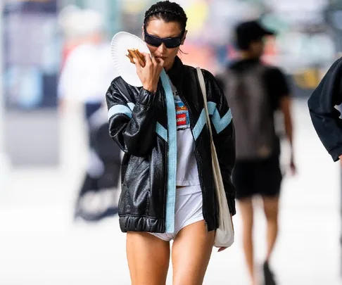 Woman in black jacket, white shorts, and sunglasses walking outdoors, holding drink and wearing a tote bag.