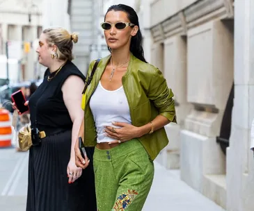 Woman in green jacket and white top with sunglasses walking on city sidewalk during NYFW SS23.