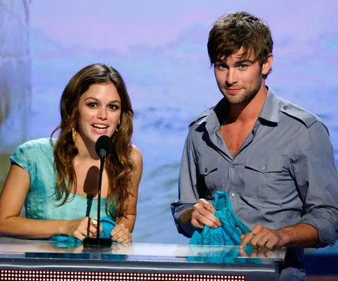 Rachel Bilson and Chace Crawford stand at a podium on stage, both smiling, against a beach-themed backdrop.