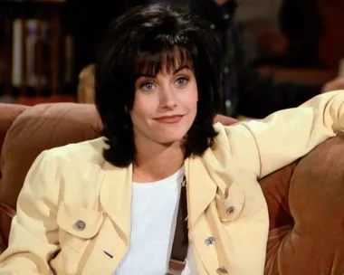 Monica from Friends with a classic '90s layered haircut and bangs, sitting on a couch wearing a light yellow jacket.