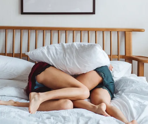 Two people in nightwear playfully hiding under a pillow on a bed.