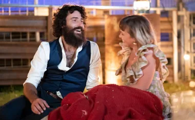 A bearded man and a woman laugh together, sitting outdoors under warm lighting, surrounded by blankets and a fence.