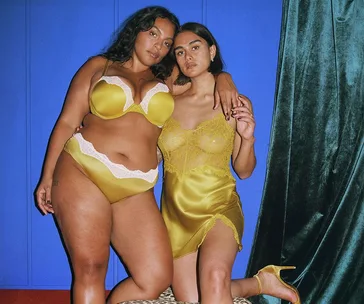 Two women in yellow lingerie pose together against a blue background, one standing and the other kneeling.