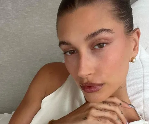 A person posing with minimal makeup, gold earrings, and tattoos on fingers, wearing a white top.