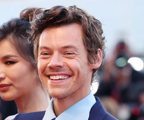 Two people smiling on a red carpet, one with curly hair and blue suit, lights and cameras in the background.
