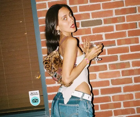 Woman in jeans and tank top holding a wine glass, standing against a brick wall with a leopard print bag.