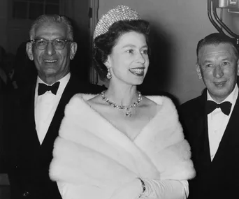 Queen Elizabeth II wearing a tiara and fur stole, smiling at an event, with two men in tuxedos beside her.