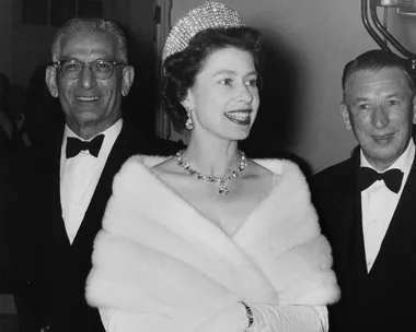 Queen Elizabeth II wearing a tiara and fur stole, smiling at an event, with two men in tuxedos beside her.