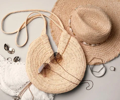 Straw hat and bag, sunglasses, jewelry, and white fabric laid out stylishly on a neutral background.