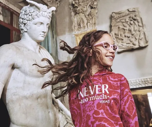 A girl in a pink Versace sweater and glasses smiles next to a marble statue.