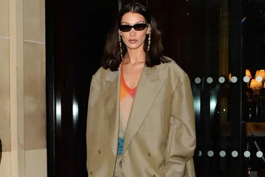 A person wearing oversized fashion with sunglasses and earrings stands in front of a building entrance at night.