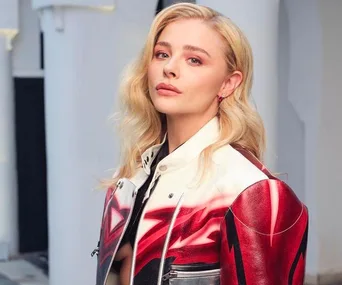 Person with long blonde hair, wearing a red and white jacket, looking at the camera against a neutral background.