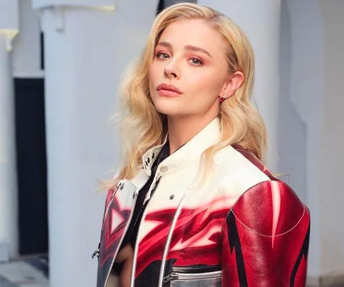 Person with long blonde hair, wearing a red and white jacket, looking at the camera against a neutral background.