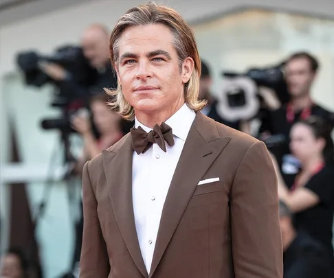 Chris Pine at the "Don't Worry Darling" premiere, wearing a brown suit and bow tie, with photographers in the background.