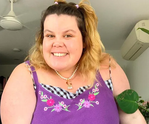 Smiling person with light hair in a purple floral top. Background includes a ceiling fan and plant.