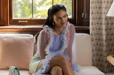 A woman in a pastel ruffled dress sits on a couch, smiling softly with a window and greenery behind her.