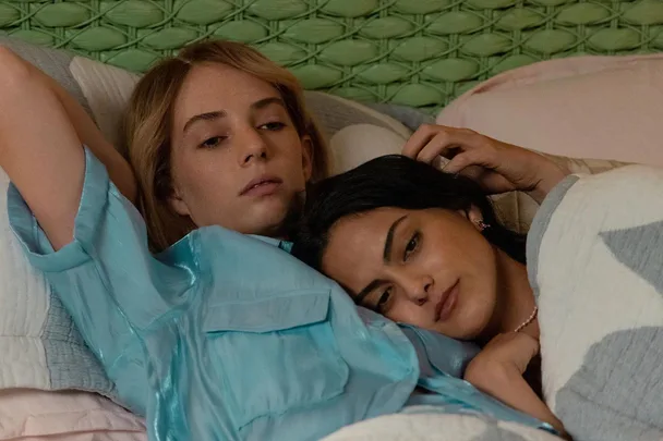 Two characters from "Do Revenge" lie in bed together, one with light hair in a blue top, the other with dark hair.