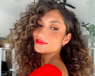 Woman with curly hair and red lipstick wearing a red top, looking over her shoulder in a bright room.