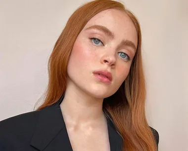 A person with long red hair and blue eyes, wearing a black blazer, against a plain background.