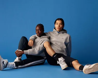 Two models seated against a blue background wearing gray Nike hoodies and sneakers, promoting sustainable fashion.