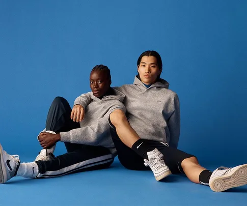 Two models seated against a blue background wearing gray Nike hoodies and sneakers, promoting sustainable fashion.