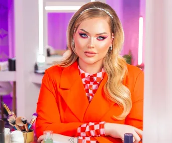 Makeup influencer with bold pink eyeshadow, wearing an orange outfit, sitting at a vanity with makeup tools.