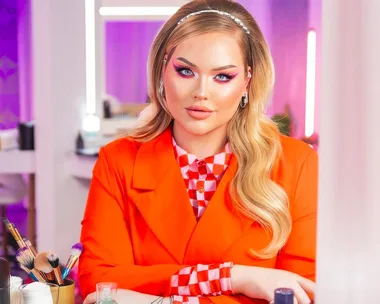 Makeup influencer with bold pink eyeshadow, wearing an orange outfit, sitting at a vanity with makeup tools.