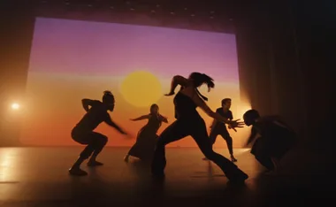 Dancers performing energetically against a backdrop of a vibrant sunset on stage, silhouetted by warm lighting.