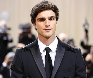 Young man in a formal black suit with embellishments, posing at a fashion event with photographers in the background.