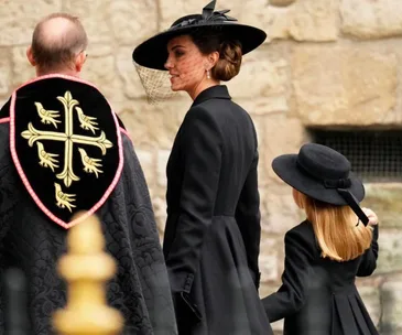 A woman and a child in black formal attire with hats stand beside a clergy member in a ceremonial robe at a stone structure.