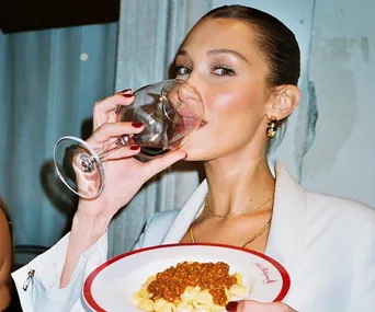 A person in a white outfit sips red wine while holding a pasta dish topped with sauce, smiling at the camera.