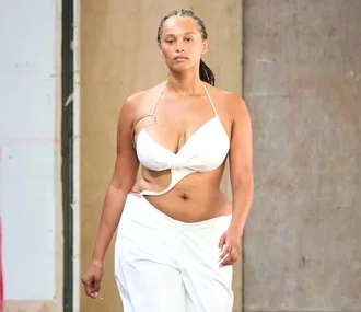 Model in a white, cut-out top and pants walks runway against a plain background.