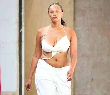 Model in a white, cut-out top and pants walks runway against a plain background.