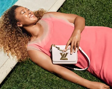 Person in pink dress lying on grass, smiling, holding a white Louis Vuitton purse near a pool.