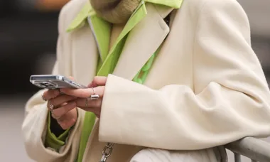 Person holding a smartphone, wearing a cream coat and green shirt, visible rings on fingers.