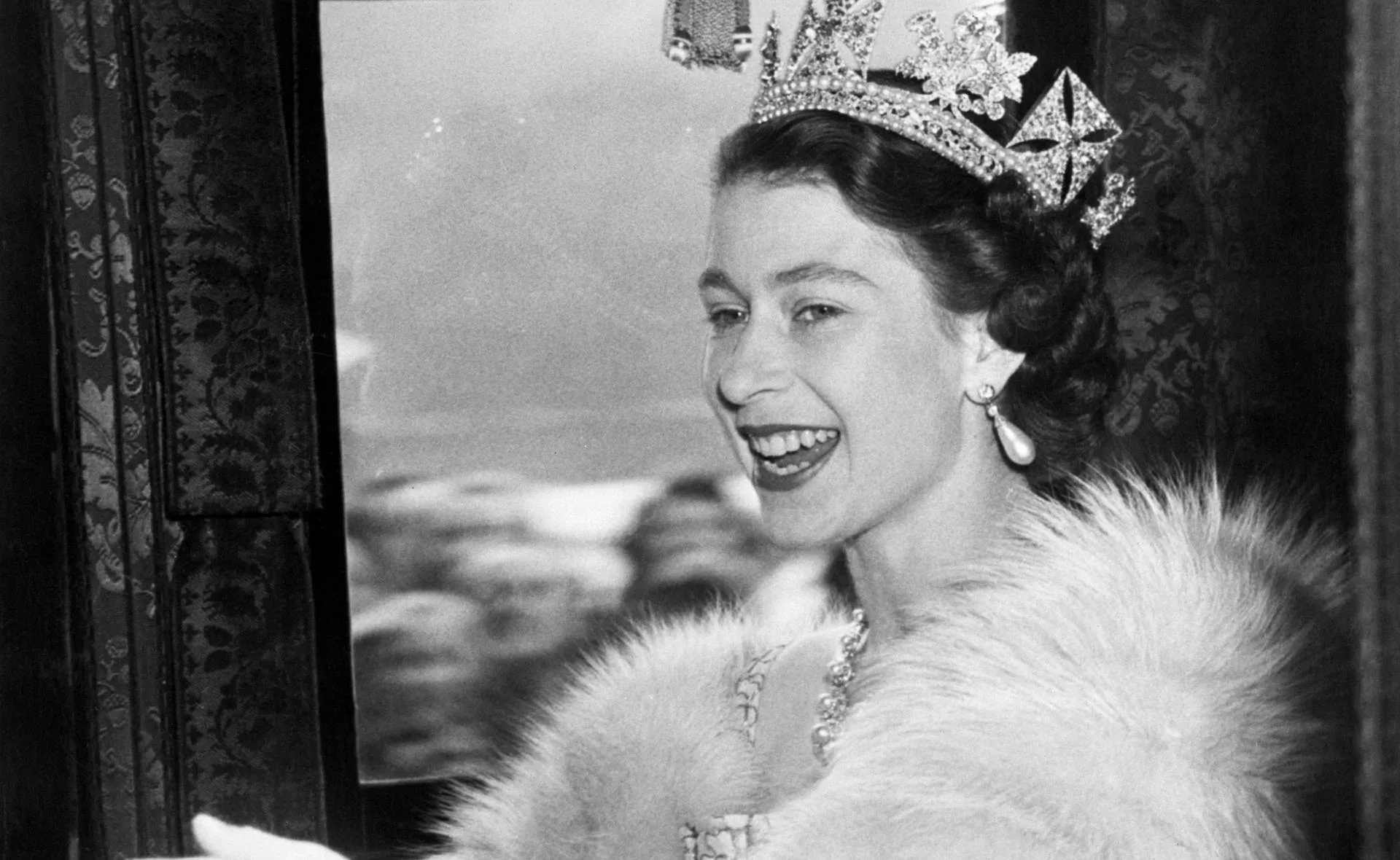 A smiling woman in a tiara and pearl earrings, wearing a fur stole, looks out of a window.