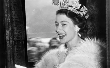 A smiling woman in a tiara and pearl earrings, wearing a fur stole, looks out of a window.