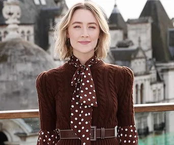 Person in a brown cable-knit sweater with polka dot scarf, standing in front of urban scenery with historic buildings.